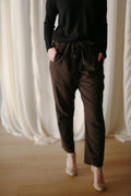 A person wearing a black long-sleeve top and Cashmere Unisex Pants | Brown with hands in pockets stands on a wooden floor. The background features sheer white curtains. Only the person's torso and legs are visible, giving a minimalist, unisex appeal to the outfit.