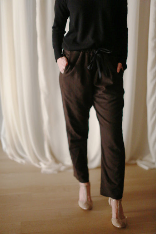 A person wearing a black long-sleeve top and Cashmere Unisex Pants | Brown with hands in pockets stands on a wooden floor. The background features sheer white curtains. Only the person's torso and legs are visible, giving a minimalist, unisex appeal to the outfit.