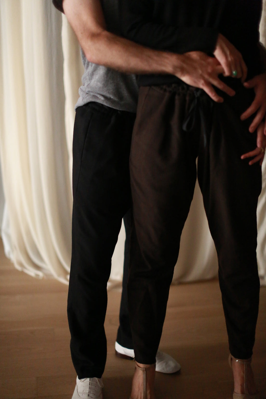 A person with their arms around another, both dressed in unisex casual clothing, standing in front of a light-colored curtain. The front person's head is not visible, and the rear person's face is partially out of frame. The atmosphere appears intimate, accentuated by their cozy Cashmere Unisex Pants | Brown.