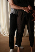 A person in a gray shirt hugs another person in a unisex black shirt and **Cashmere Unisex Pants | Black** from behind. They stand on a wooden floor with a white curtain in the background.