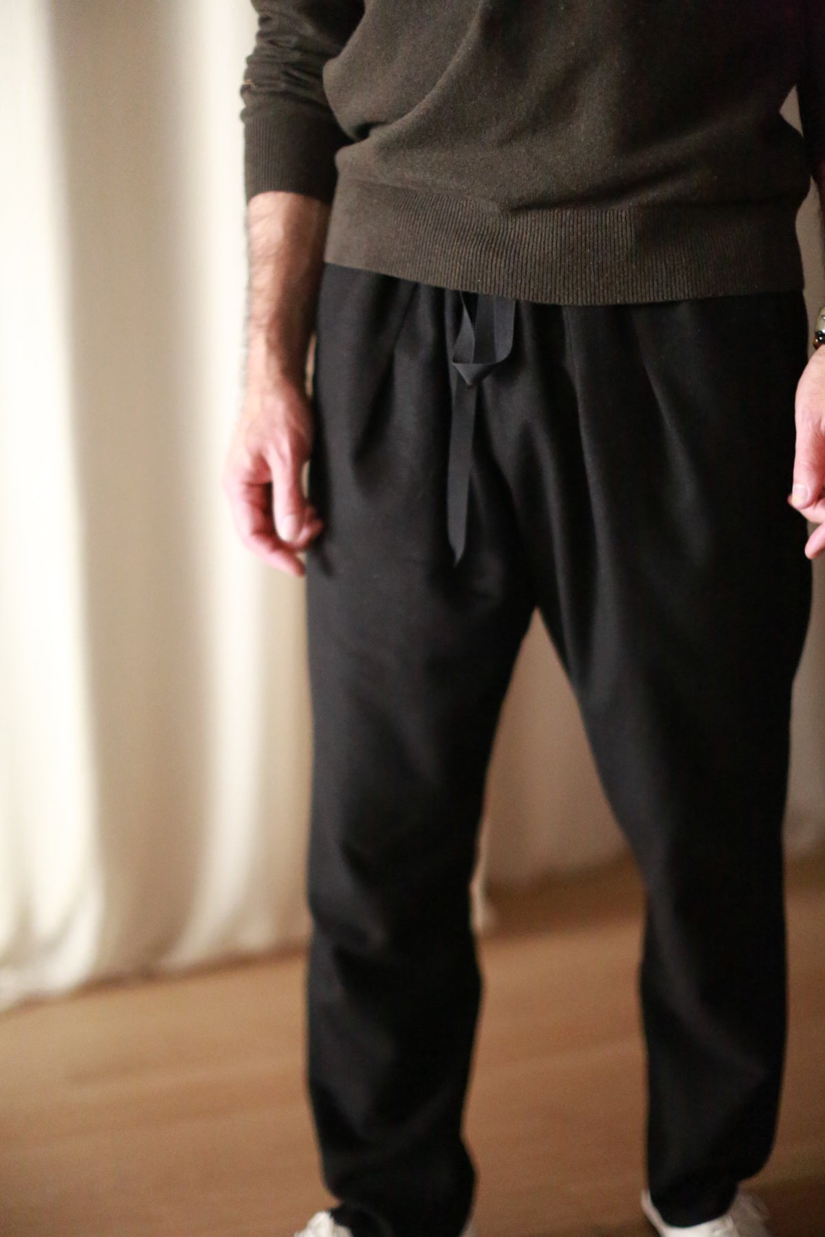 A person is shown from shoulders to feet, wearing a dark long-sleeve sweater and Cashmere Unisex Pants | Black with a silk ribbon drawstring waist. The background features white curtains and a wooden floor. The person's head and feet are not visible in the image.