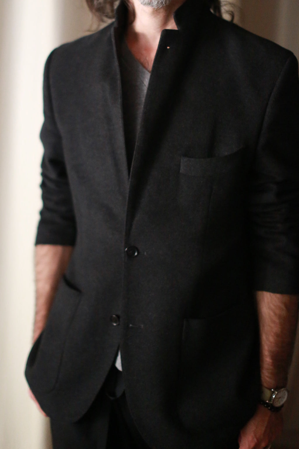 A man is depicted from mid-face to upper thighs wearing a Men's Cashmere Blazer in black over a gray shirt. This European fit blazer, meticulously crafted, boasts a high collar and two buttons. He has long hair and a beard, with his left arm relaxed by his side, accessorized with a watch.