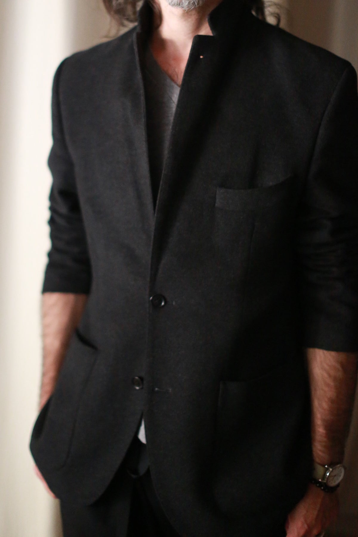 A man is depicted from mid-face to upper thighs wearing a Men's Cashmere Blazer in black over a gray shirt. This European fit blazer, meticulously crafted, boasts a high collar and two buttons. He has long hair and a beard, with his left arm relaxed by his side, accessorized with a watch.