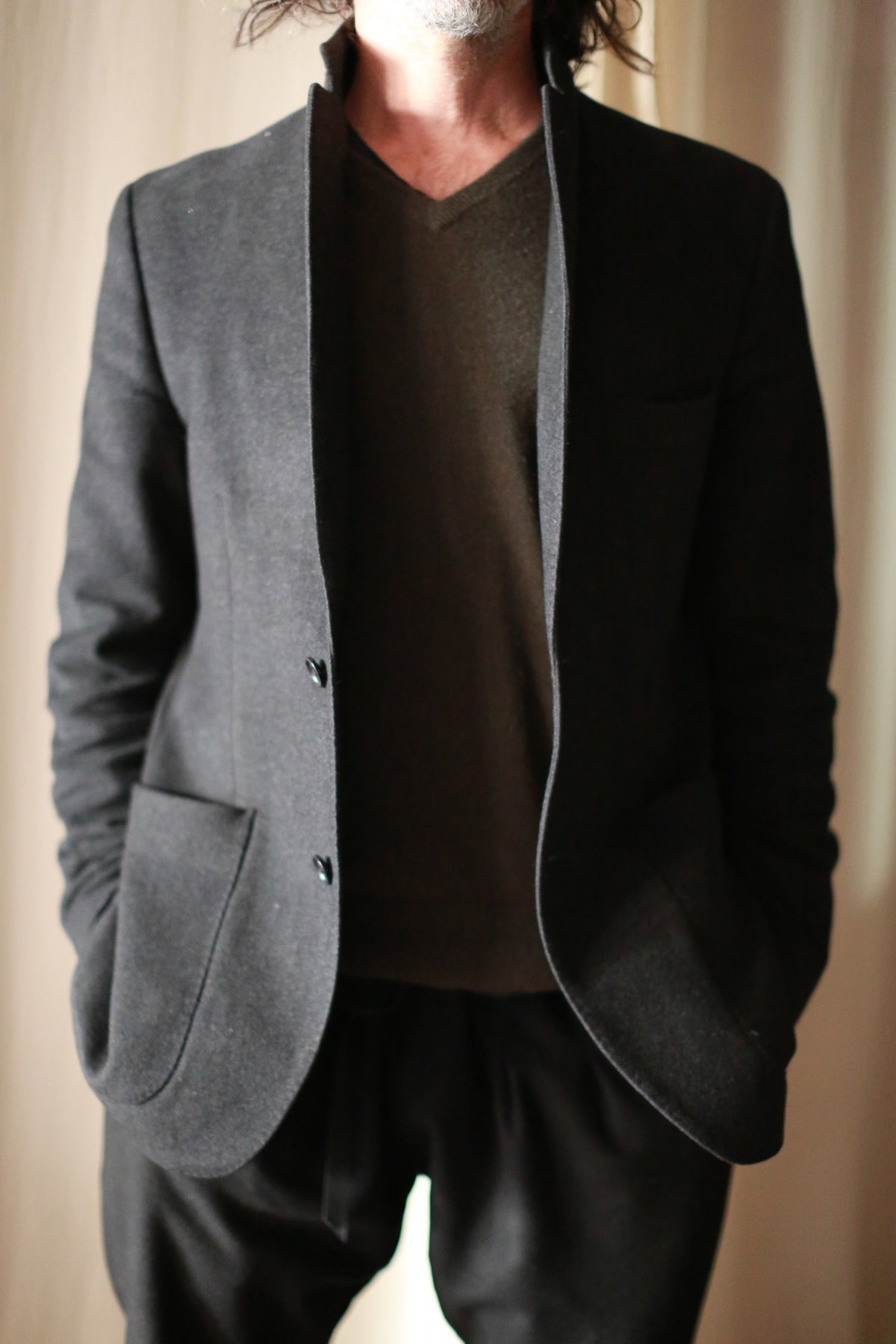 A person with long hair is seen wearing the Men's Cashmere Blazer in a dark shade, layered over a similarly dark V-neck shirt, tailored in a European fit. With their hands tucked into the blazer's pockets, they stand against a neutral background, with their face not visible.