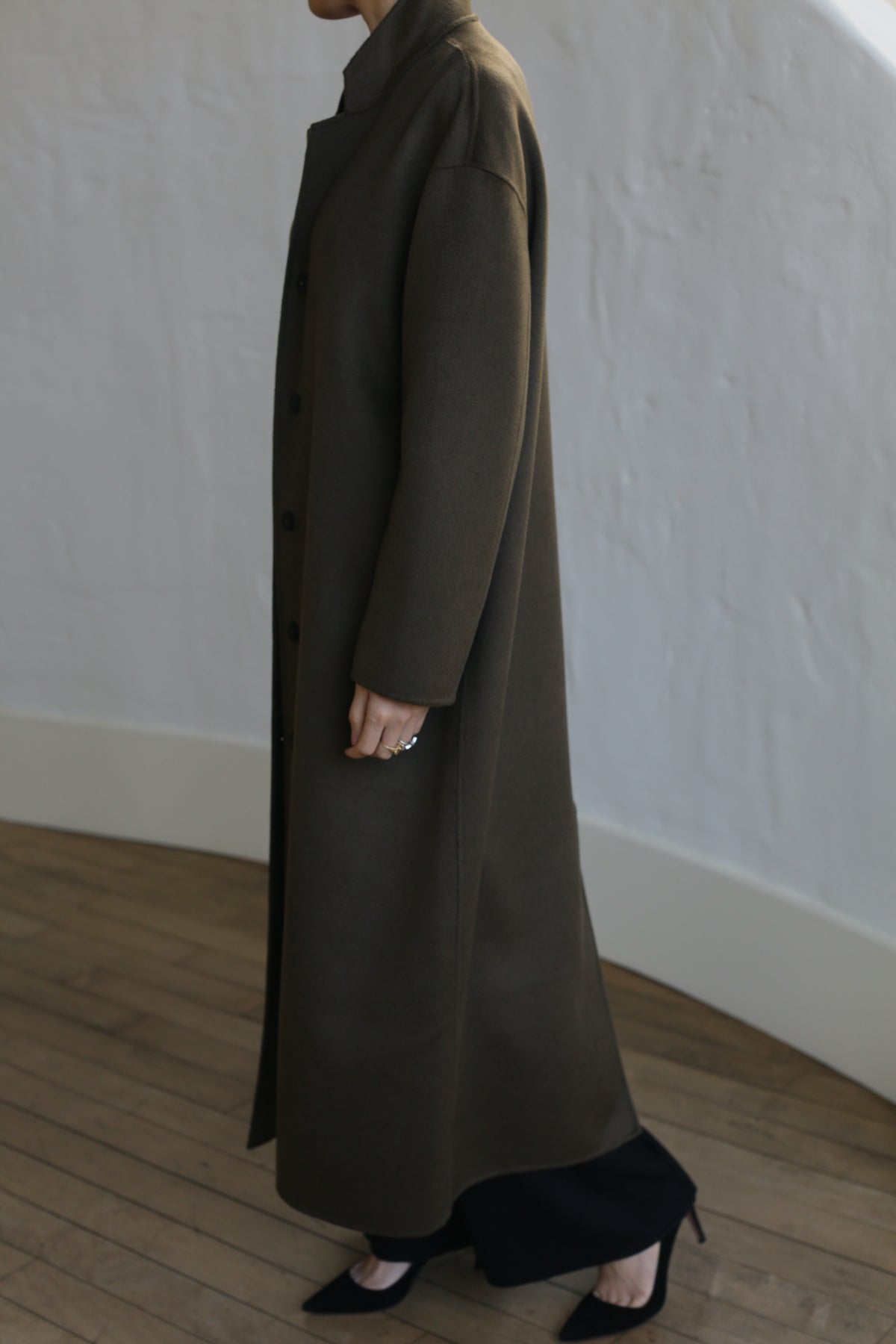 Dressed in the Bamford Long Double-face Cashmere coat in Olive, a person stands stylishly indoors against a light-textured wall. Viewed from the side, their ring-adorned hand adds elegance to the scene, complemented by the wooden floor beneath.
