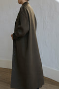 A person stands with their back to the camera, wearing a chic Bamford Long Double-face Cashmere coat in olive. The coat, crafted from luxurious material, features a simple design falling below the knees. The background shows a white textured wall and wooden floor.