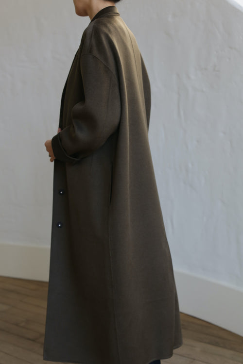 A person stands with their back to the camera, wearing a chic Bamford Long Double-face Cashmere coat in olive. The coat, crafted from luxurious material, features a simple design falling below the knees. The background shows a white textured wall and wooden floor.