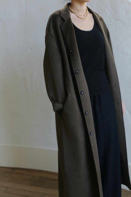 A person stands against a light wall, wearing an olive Bamford Long Double-face Cashmere coat. The luxurious coat, partially buttoned, overlays a black top and trousers. A simple necklace adds elegance, while the image emphasizes the clothing over facial details.