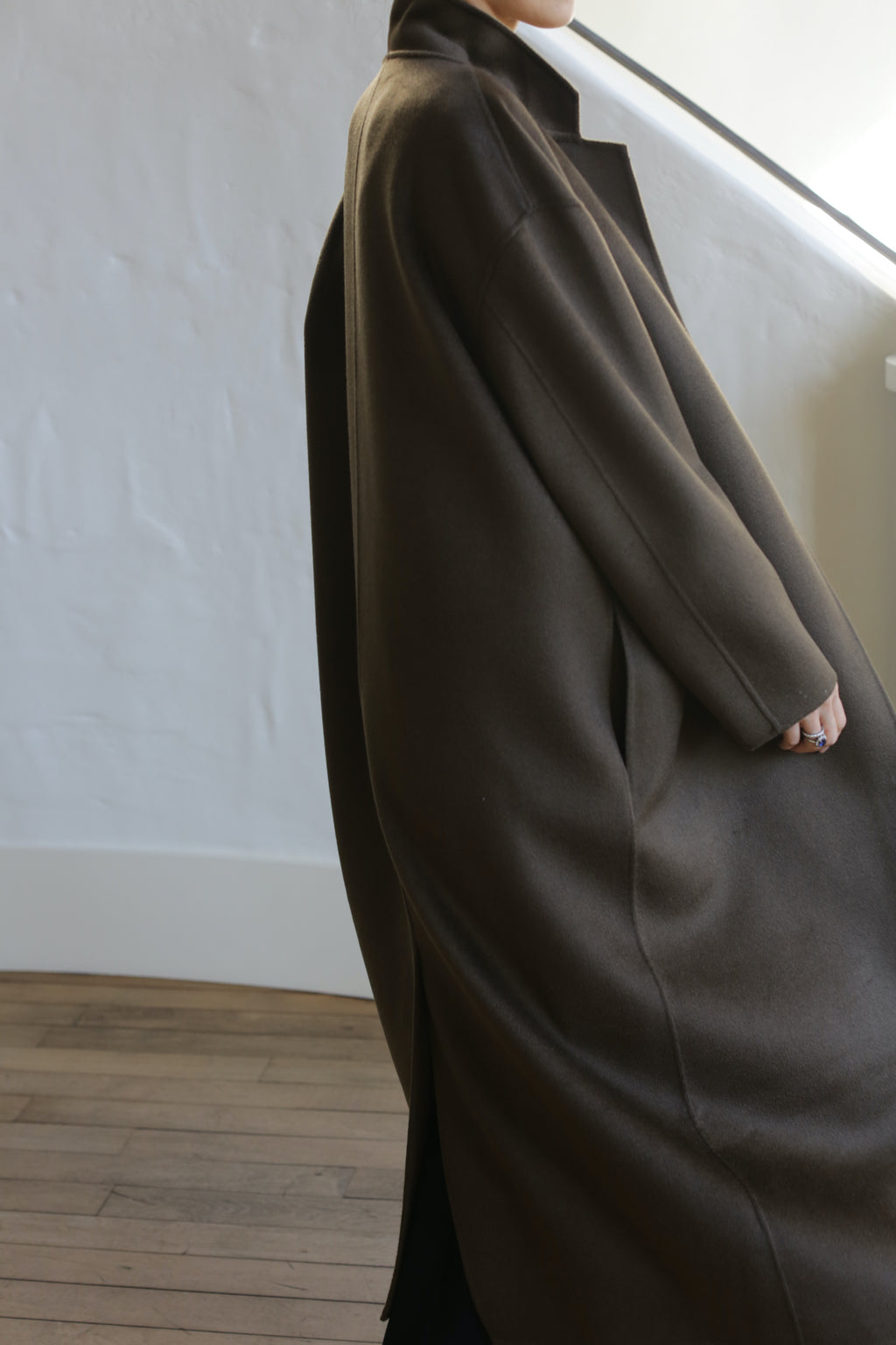 An individual wearing the Bamford Long Double-face Cashmere coat in Olive stands indoors near a white wall and stairs. The oversized coat enhances the minimalist aesthetic, accentuated by wooden flooring, with their face hidden from view.