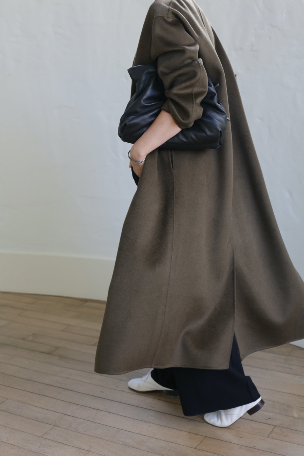 A person in a chic Bamford Long Double-face Cashmere coat in olive and black pants strolls on wooden flooring with a large black clutch. Their white shoes contrast elegantly with the light-colored wall in the background.