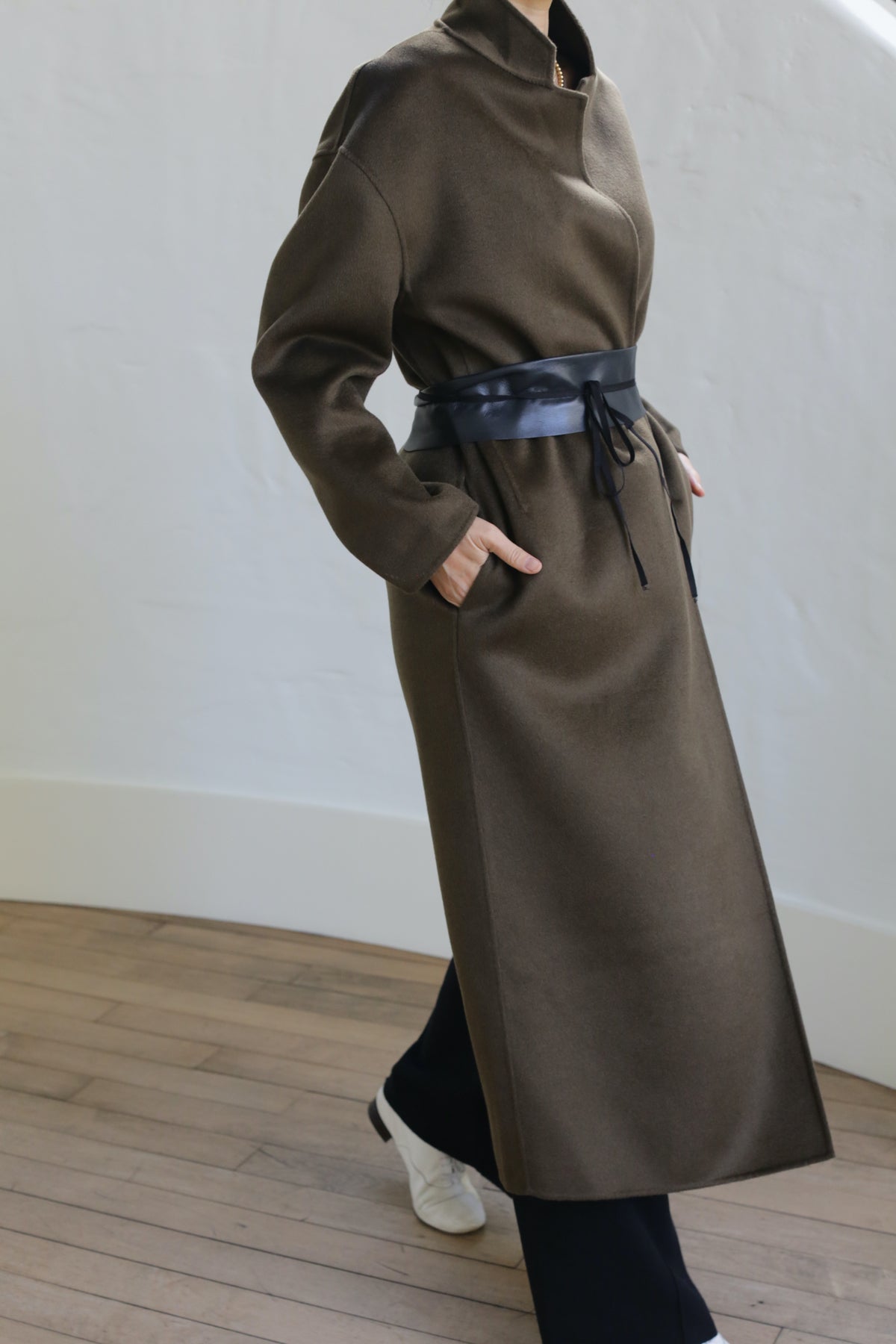 Indoors on a wooden floor against a plain white wall, the individual wears a Bamford Long Double-face Cashmere coat in olive. The stylish long coat with a black belt pairs perfectly with black pants and white shoes as their hands rest casually in the pockets.