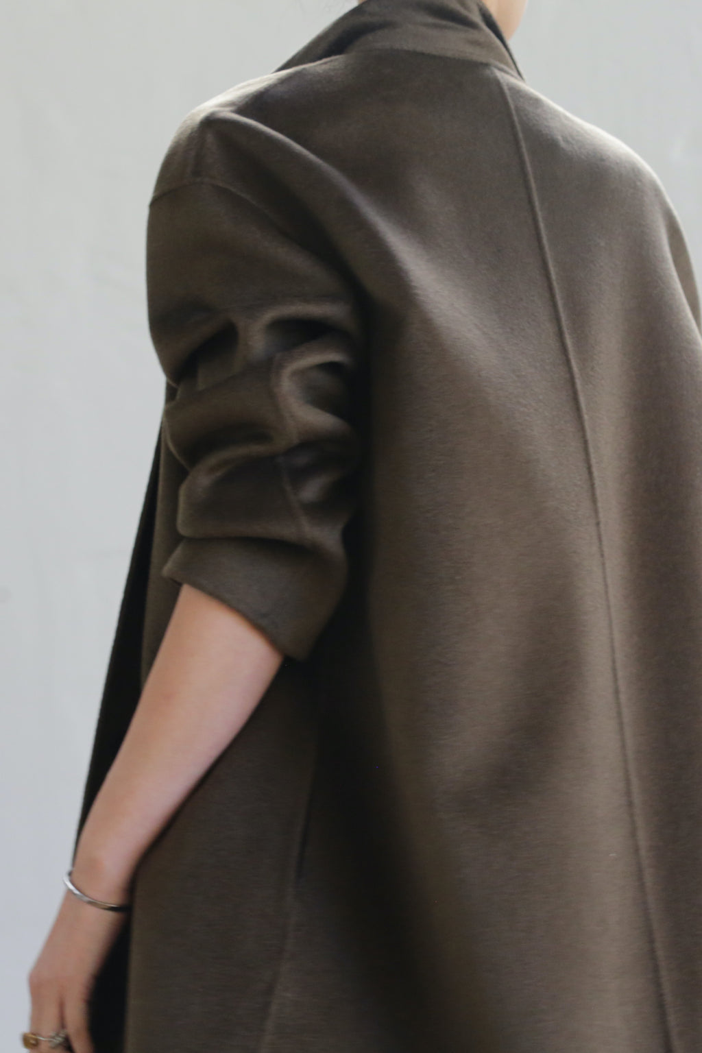 Someone wearing the Bamford Long Double-face Cashmere coat in olive, featuring a chic wide sleeve and luxurious drape, stands with their arm slightly bent. The elegant back creates a sophisticated silhouette against a plain background.