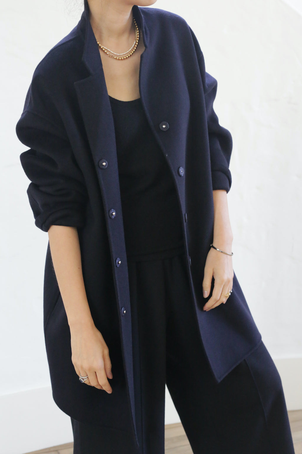 A person stands against a light background wearing the Bamford Double-face Cashmere in navy over a black top and cashmere trousers, accessorized with a gold necklace and bracelet.
