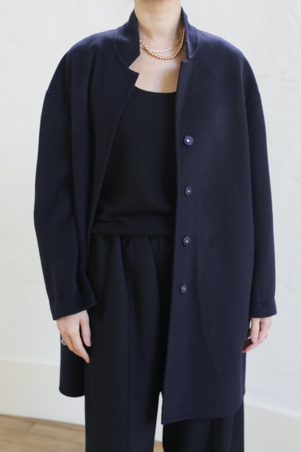 Wearing the Bamford Double-face Cashmere coat in navy over a black top and pants, a person stands against a light background. Their head is partially out of frame, and they accessorize with a gold chain necklace.