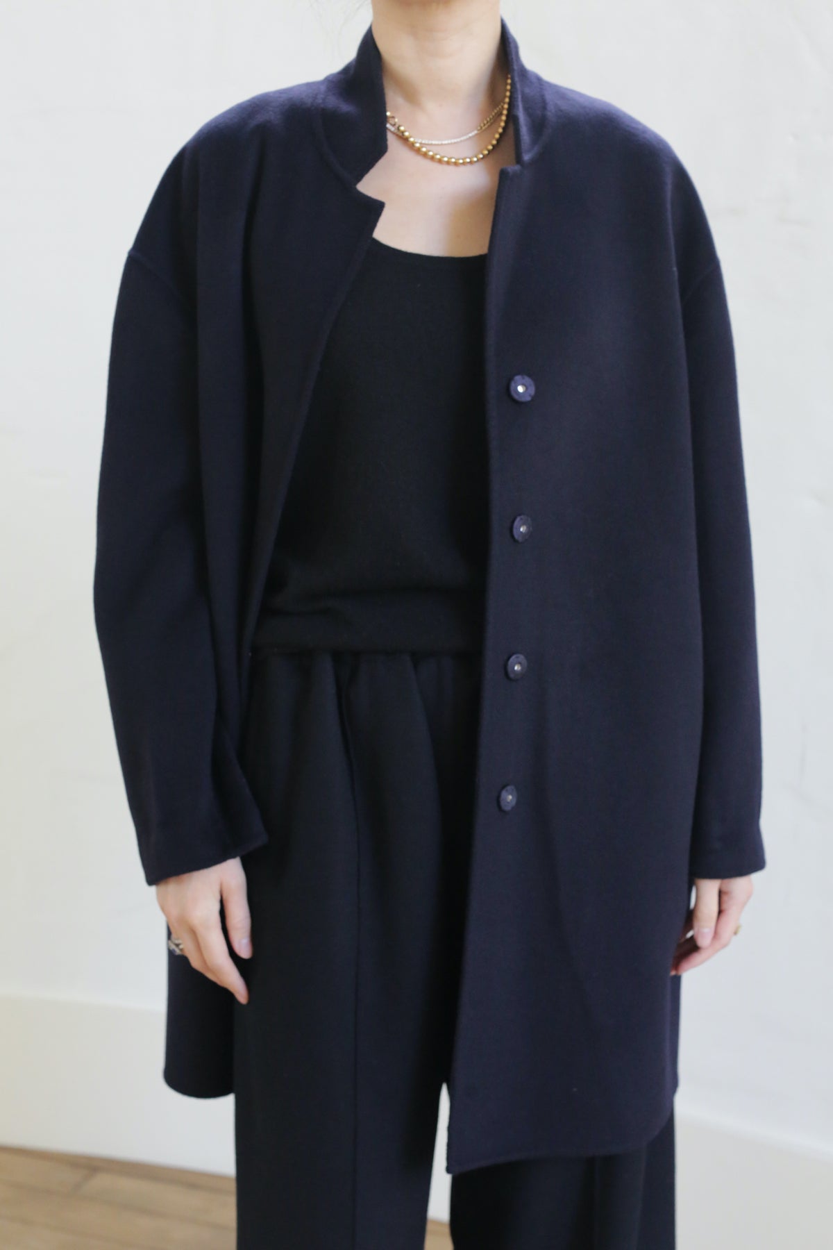 Wearing the Bamford Double-face Cashmere coat in navy over a black top and pants, a person stands against a light background. Their head is partially out of frame, and they accessorize with a gold chain necklace.