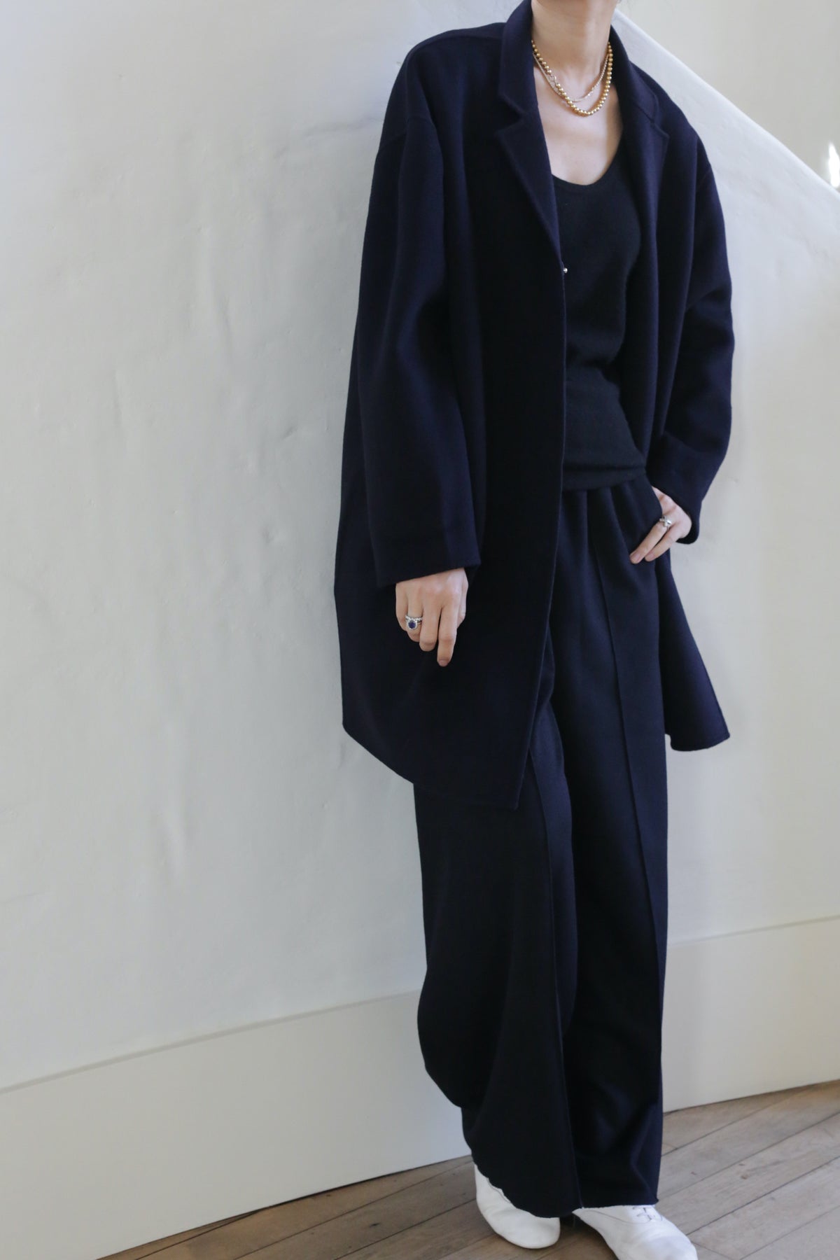 Against a white textured wall, a person in a loose black oversized blazer, black top, and Bamford Double-face Cashmere wide-leg trousers in navy stands with white shoes and layered gold necklaces, one hand in pocket—their face obscured.