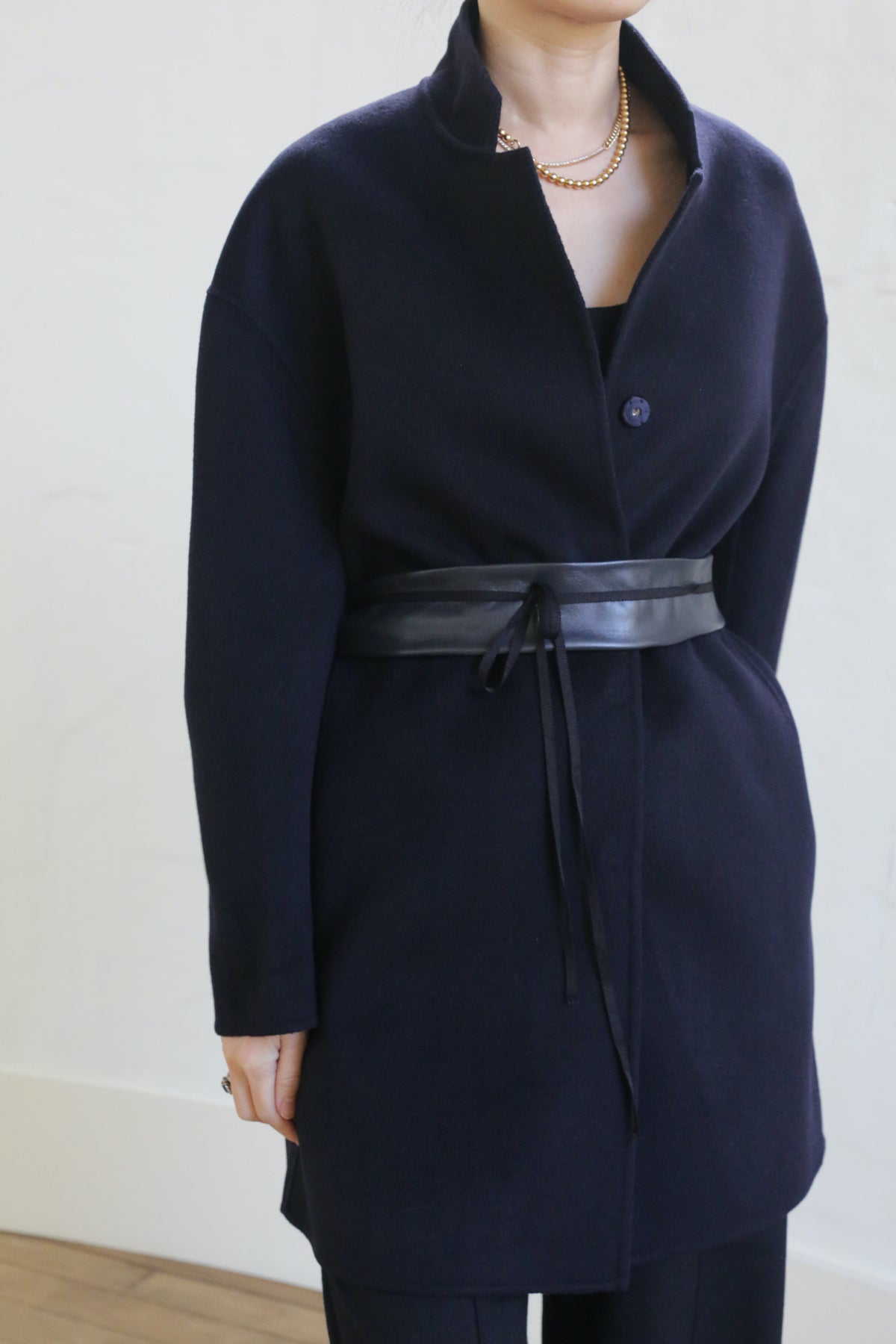 A person wearing the Bamford Double-face Cashmere coat in navy, cinched with a black leather belt, paired with a gold chain necklace and matching cashmere trousers, stands against a plain light background. The face is partially out of frame.