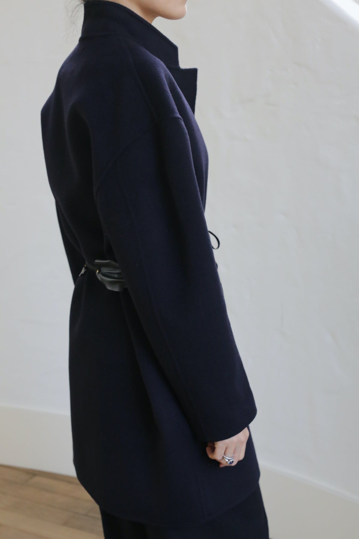 A person wearing the Bamford Double-face Cashmere coat in navy, belted, is shown from the side against a white wall. Only their upper body and part of their face are visible.