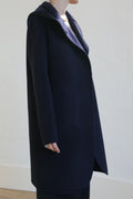 A person in the Mitchley Long Double-face Cashmere coat in navy, featuring a notched collar and handmade snap buttons, stands sideways against a light wall. The minimalist design keeps their face out of view.
