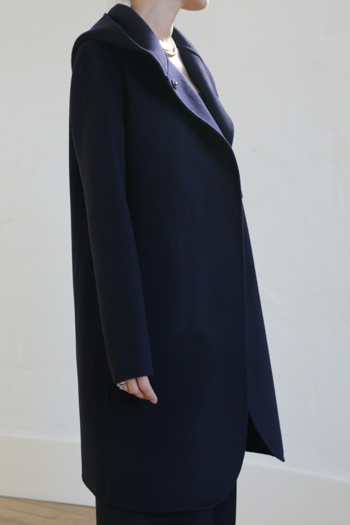 A person in the Mitchley Long Double-face Cashmere coat in navy, featuring a notched collar and handmade snap buttons, stands sideways against a light wall. The minimalist design keeps their face out of view.