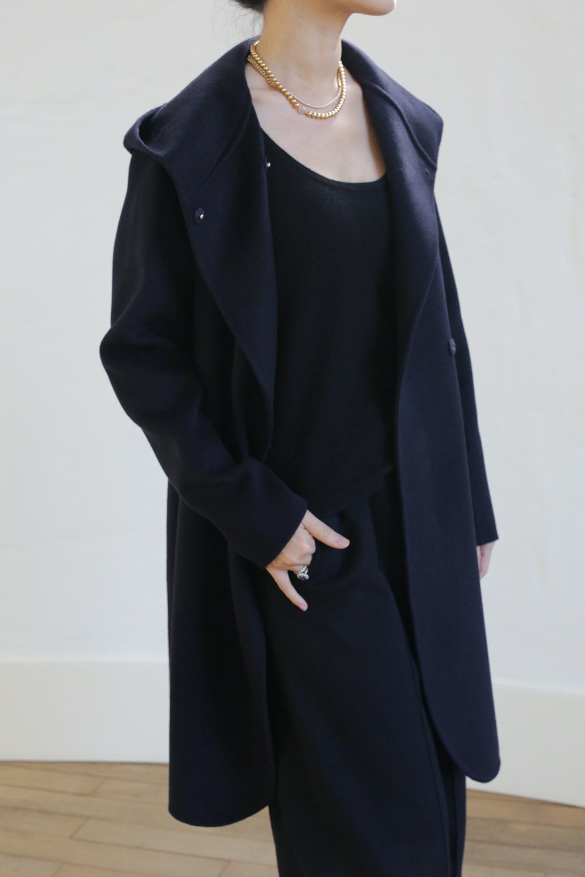 A woman wears the Mitchley Long Double-face Cashmere coat in navy over a matching set, accessorized with a gold chain necklace. Her face is partially out of frame against a light background, with one hand in her pocket.