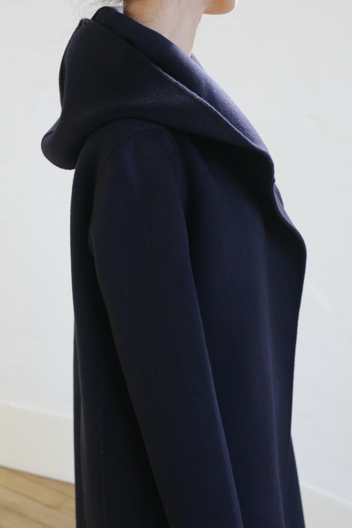 A person in the Mitchley Long Double-face Cashmere coat in navy is shown from the side, head slightly turned with the hood resting on their back against a white minimalistic background.