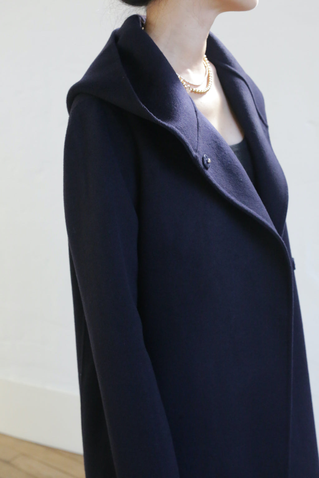 Wearing the Mitchley Long Double-face Cashmere coat in navy with a wide collar and elegant handmade snap buttons, paired with a gold necklace. The person stands indoors against a light wall, face not visible.