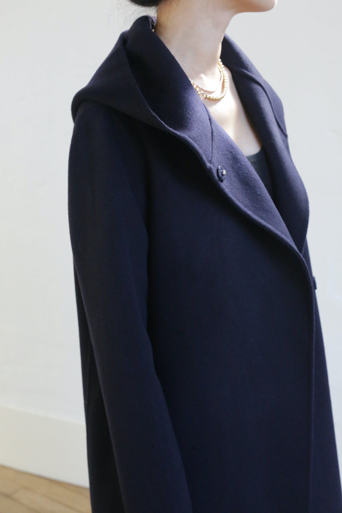 Wearing the Mitchley Long Double-face Cashmere coat in navy with a wide collar and elegant handmade snap buttons, paired with a gold necklace. The person stands indoors against a light wall, face not visible.