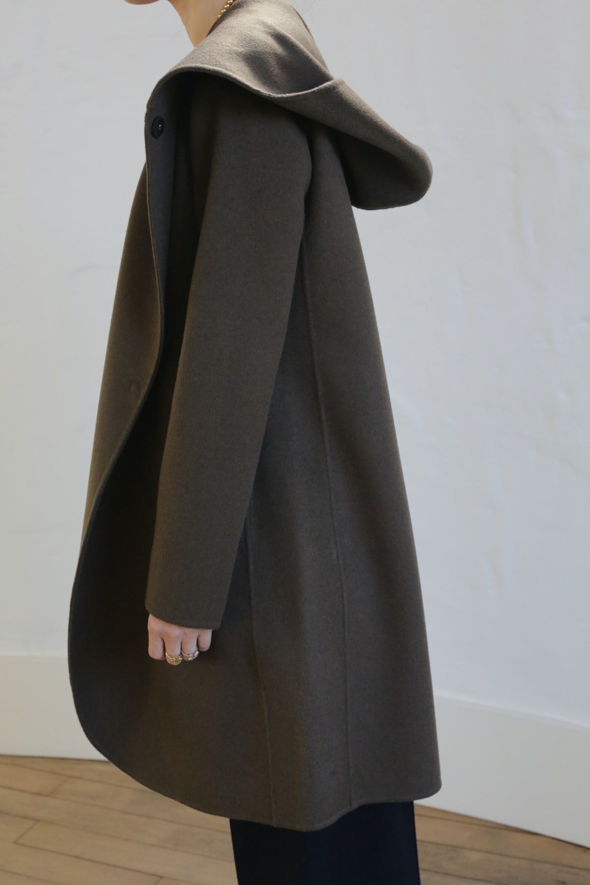 A person in an olive Mitchley Long Double-face Cashmere coat with a deep hood stands sideways. The handmade snap button subtly glints as the light-colored wall and wooden flooring form the backdrop, concealing their face from view.