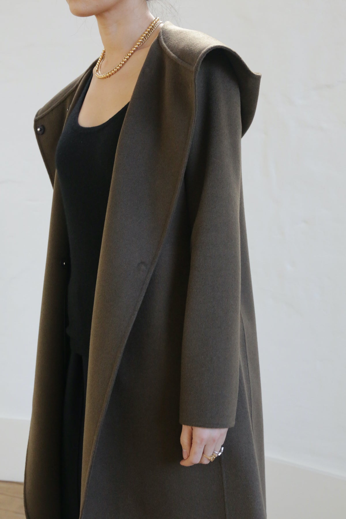 A person wearing the Mitchley Long Double-face Cashmere coat in olive with a deep hood over a black outfit stands against a light background. The coat is accessorized with handmade snap buttons, a gold necklace, and rings. Only the upper body is visible.