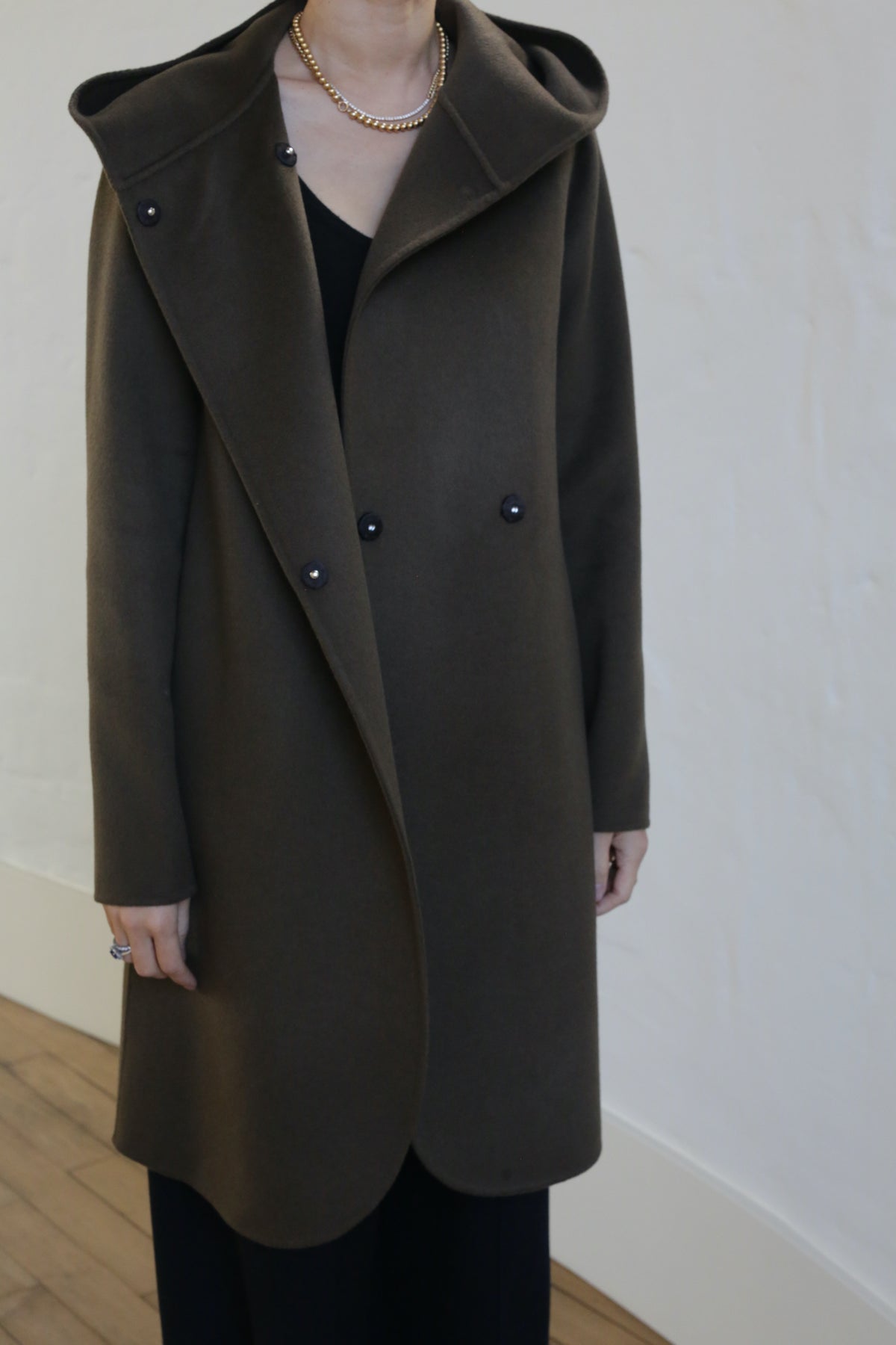 A person in a Mitchley Long Double-face Cashmere coat, olive-colored and double-breasted with handmade snap buttons and a deep hood, paired with a gold chain necklace. The coat is buttoned over sleek black clothing, standing against a light-colored wall and wooden floor.