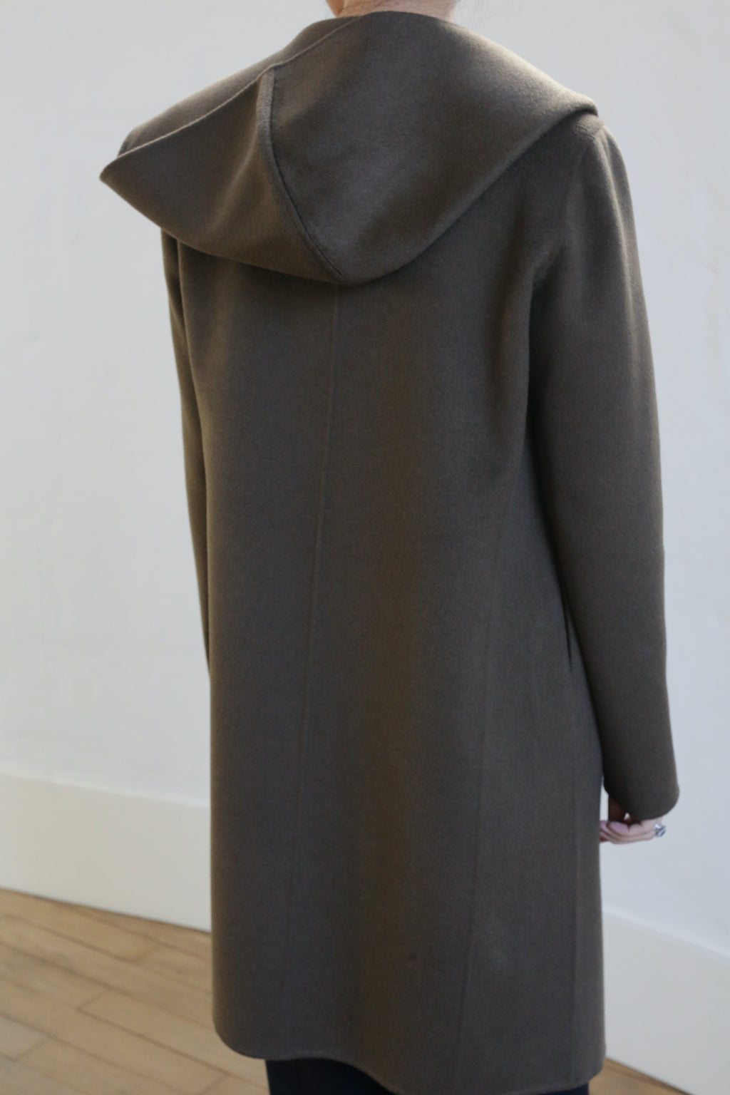 Someone in a Mitchley Long Double-face Cashmere coat in olive stands indoors, hood up. This elegant piece features handmade snap buttons and contrasts beautifully with the simple, light-colored wall and wooden floor.