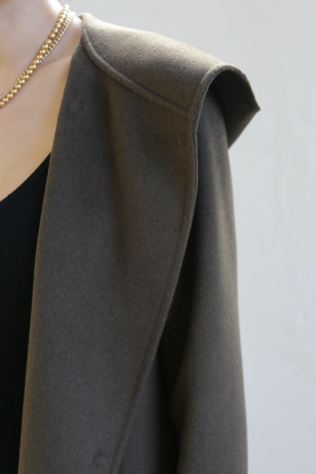 Close-up of a person wearing the Mitchley Long Double-face Cashmere coat in olive, showcasing handmade snap buttons and a gold chain necklace. The wide lapel is worn over a black top, with the image focusing on the shoulder and neckline to highlight intricate details.