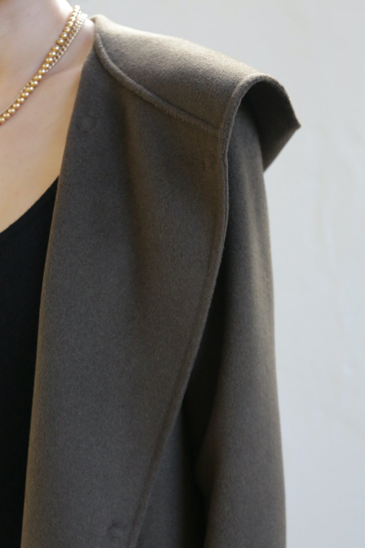 Close-up of a person wearing the Mitchley Long Double-face Cashmere coat in olive, showcasing handmade snap buttons and a gold chain necklace. The wide lapel is worn over a black top, with the image focusing on the shoulder and neckline to highlight intricate details.