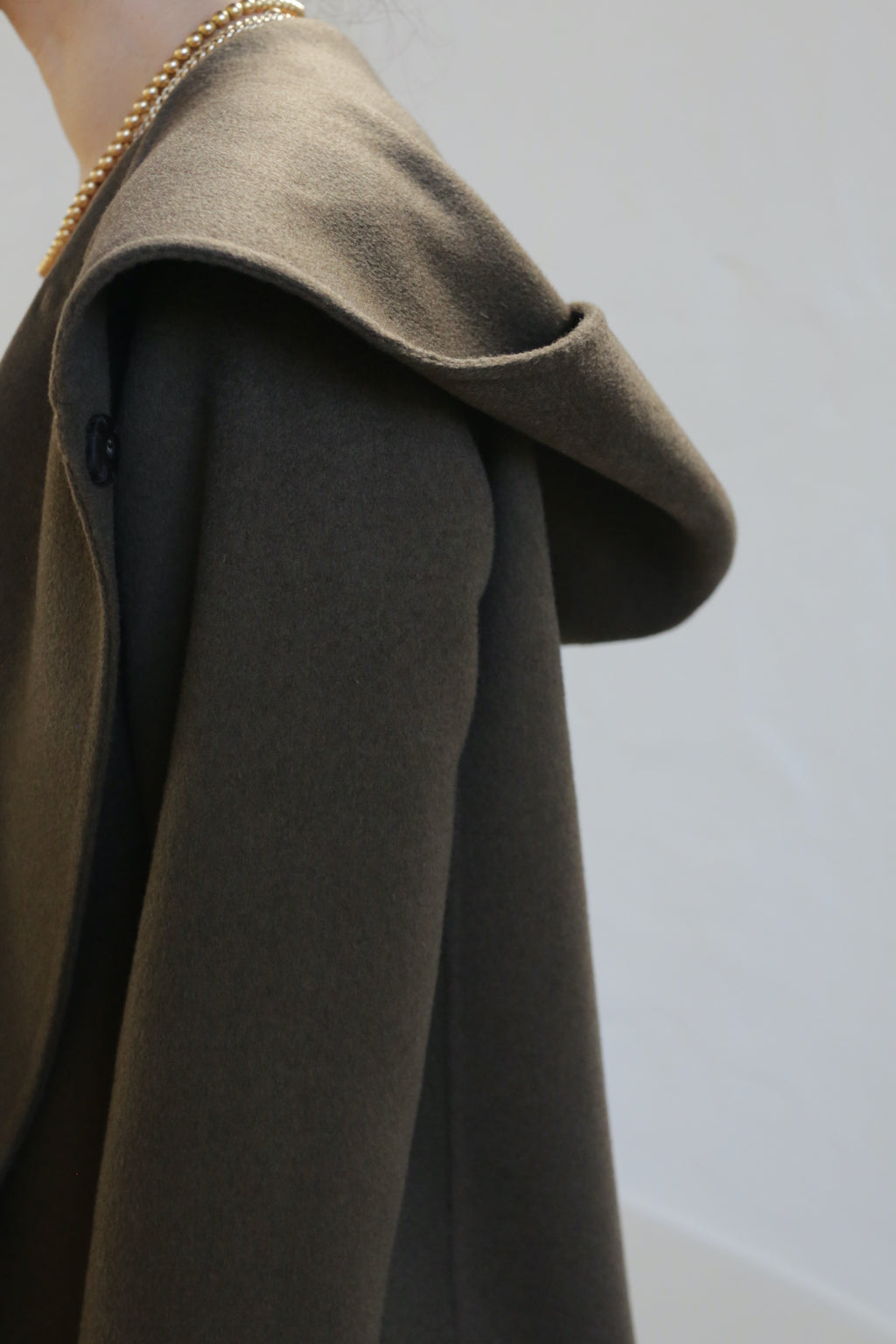 A person wears the olive Mitchley Long Double-face Cashmere coat with a deep hood, featuring handmade snap buttons and a gold chain necklace. The minimalist design stands out against a softly lit plain background.