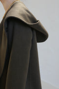 A person wears the olive Mitchley Long Double-face Cashmere coat with a deep hood, featuring handmade snap buttons and a gold chain necklace. The minimalist design stands out against a softly lit plain background.