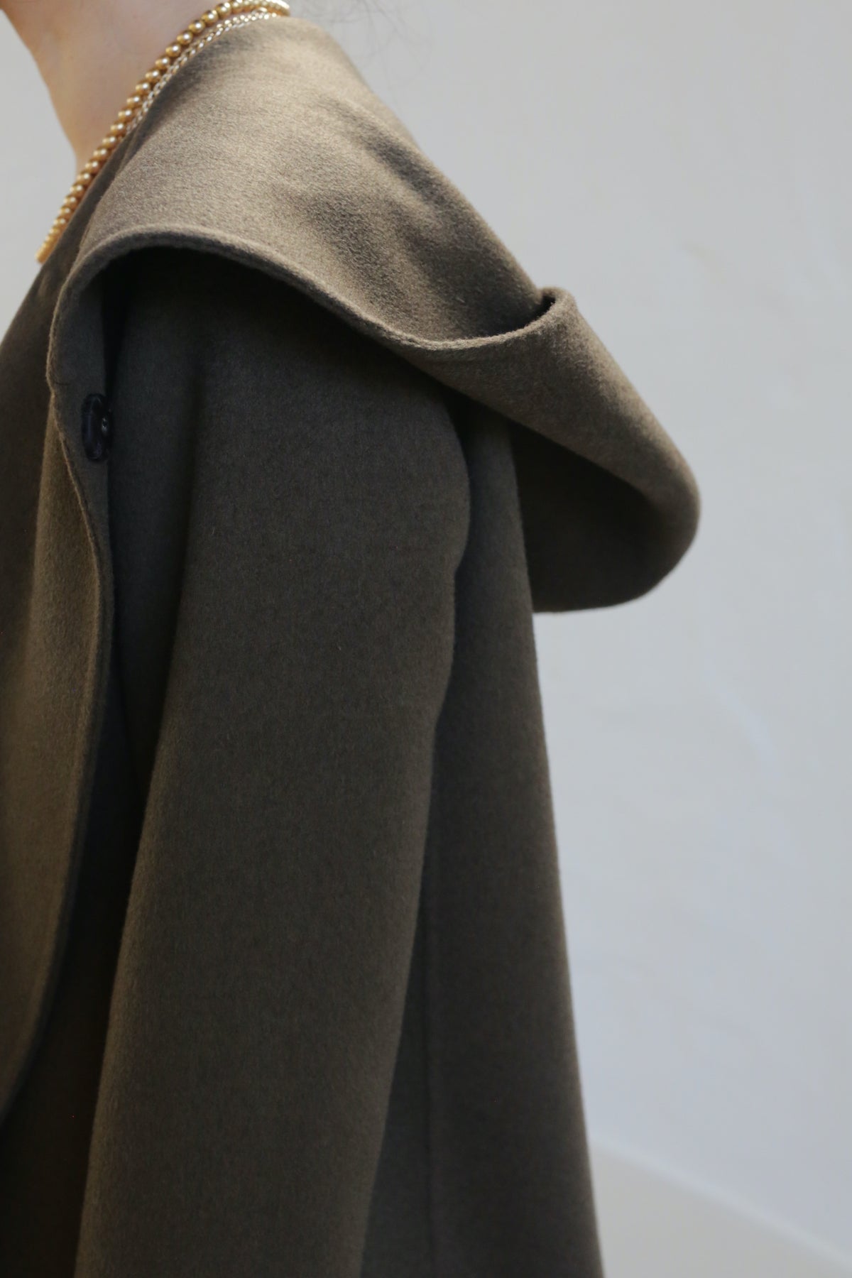 A person wears the olive Mitchley Long Double-face Cashmere coat with a deep hood, featuring handmade snap buttons and a gold chain necklace. The minimalist design stands out against a softly lit plain background.