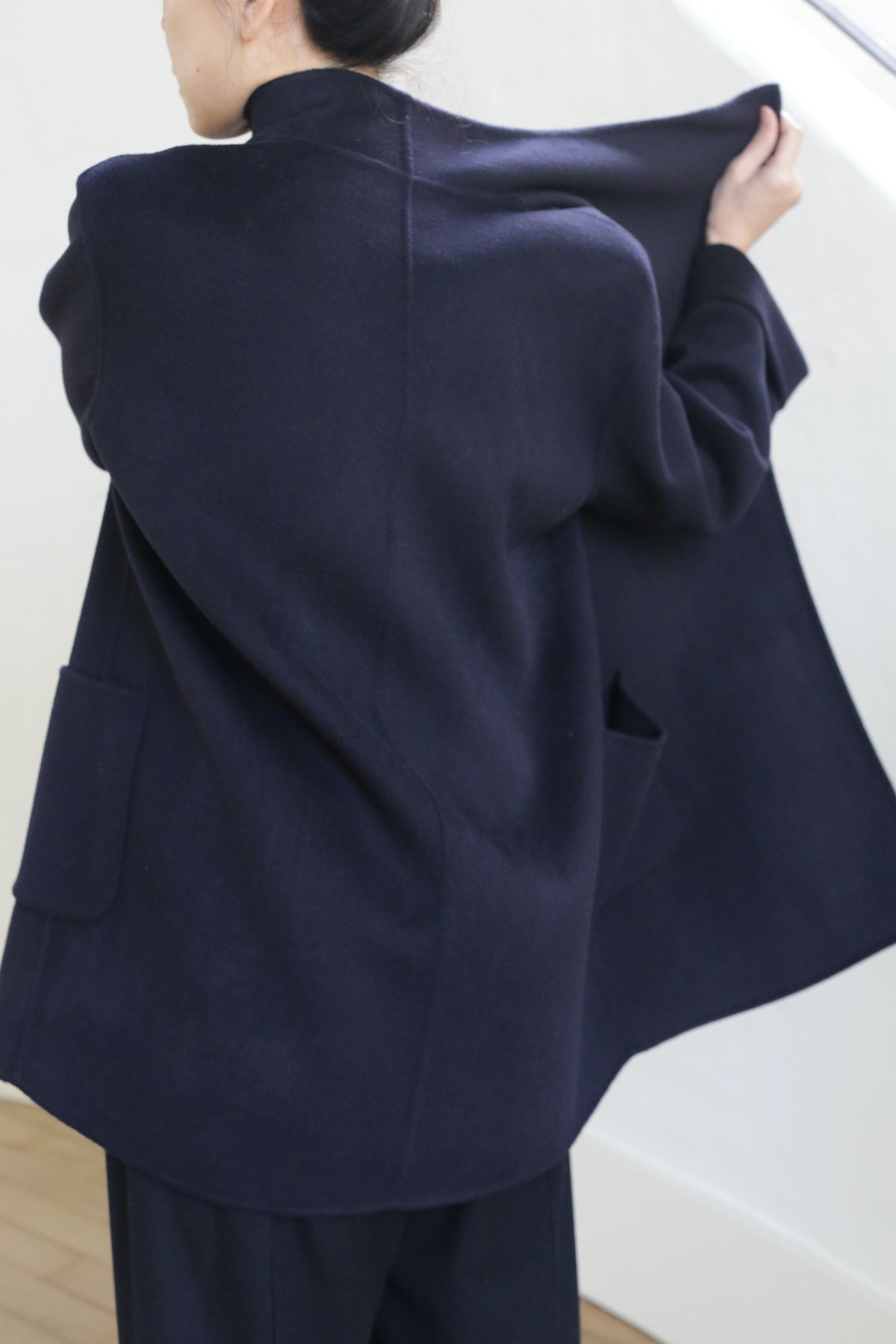 A person with dark hair in a bun is seen from behind, wearing the Winston Double-face Cashmere jacket in navy and matching pants, holding one side open near a white wall and wooden floor.