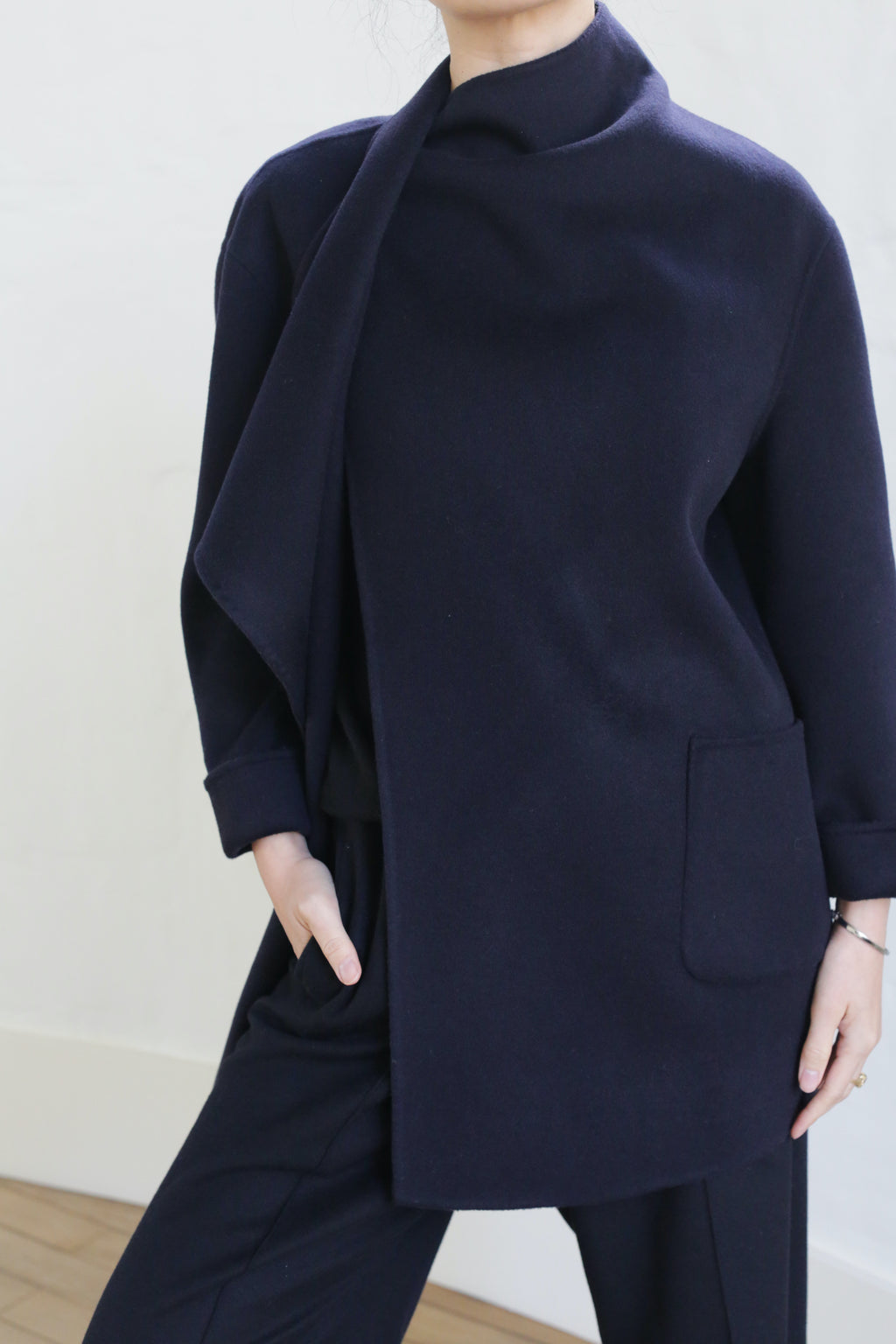 A person wearing the Winston Double-face Cashmere jacket in navy, with long sleeves, a draped high collar, and front pocket, paired with matching dark pants stands against a light background. Their face is partially out of frame.