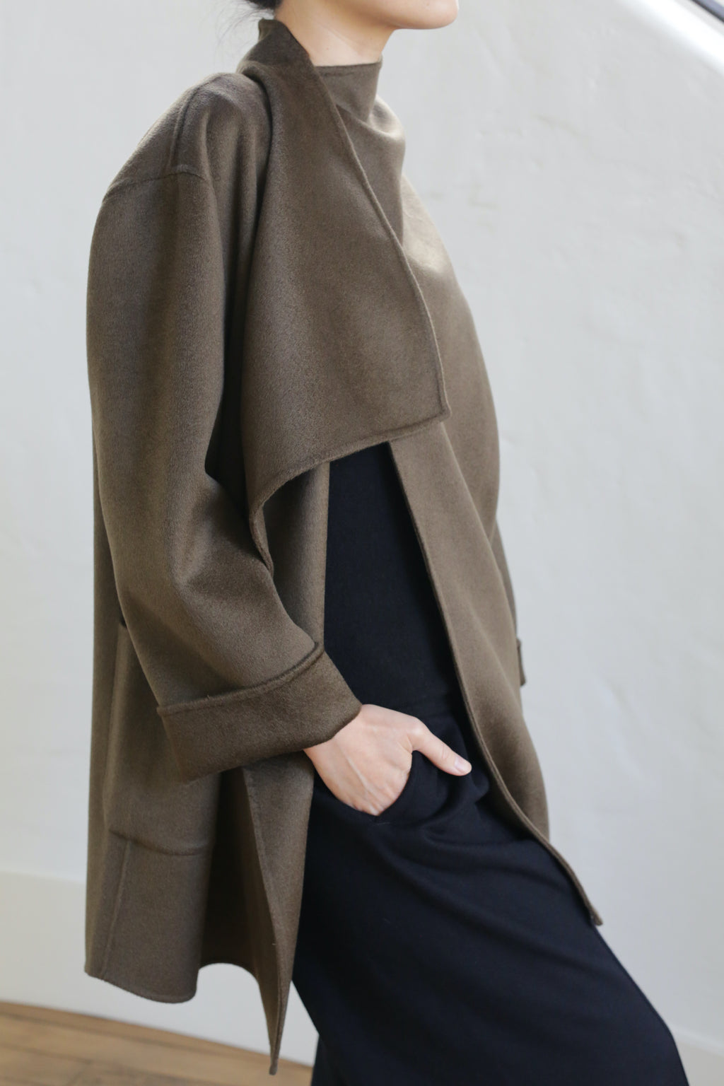 A person stands with one hand in their pocket, wearing the Winston Double-face Cashmere jacket in Olive over a long black outfit against a plain, light background. Their face is partially out of frame.