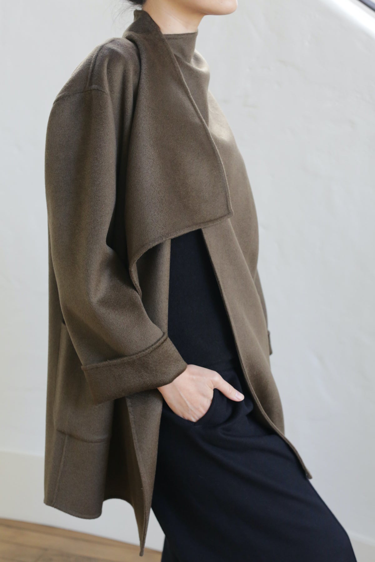 A person stands with one hand in their pocket, wearing the Winston Double-face Cashmere jacket in Olive over a long black outfit against a plain, light background. Their face is partially out of frame.