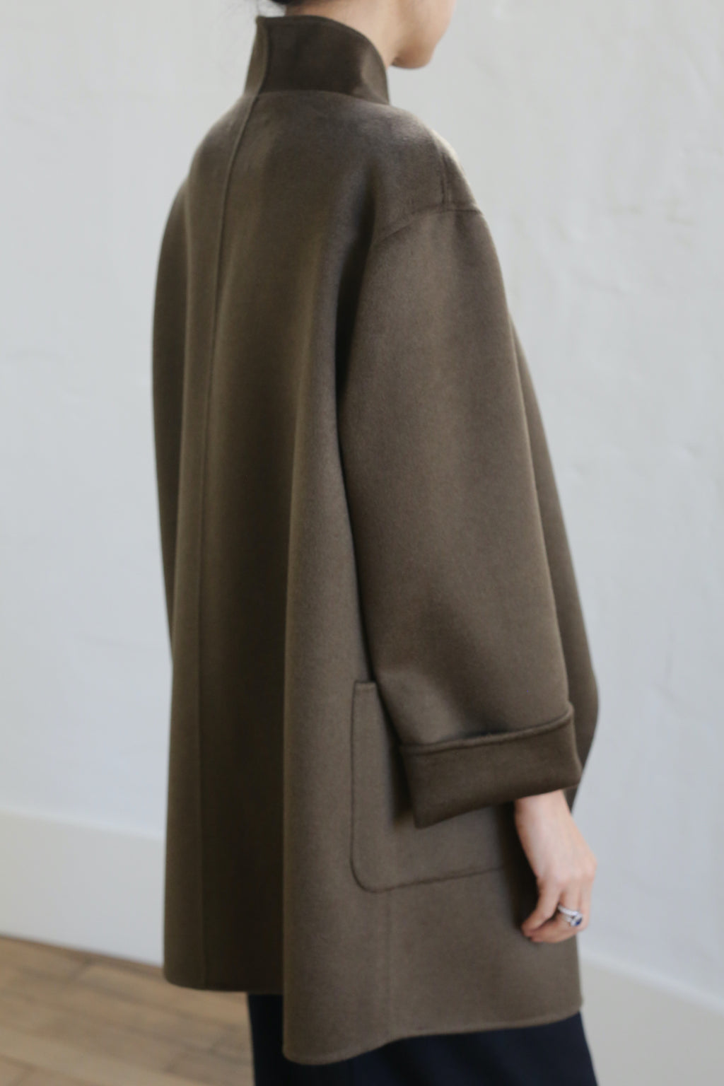 A person in the Winston Double-face Cashmere coat in olive, featuring wide sleeves and a large pocket, stands against a light wall. The loose-fitting jacket is shown from the back and side with the wearer’s hand partly visible.