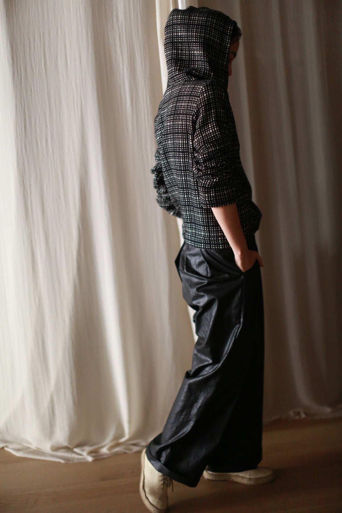 A person in the Alex Hoodie | Plaid with a silk ribbon waist and black pants stands sideways near white curtains, hands in pockets. Soft lighting highlights the texture of the outfit.