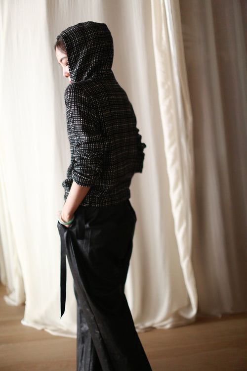 A person stands sideways wearing the Alex Hoodie | Plaid in cashmere and loose black pants, with a hand in their pocket, looking down. The soft, flowing white curtain background evokes artisan creations handcrafted in San Francisco.
