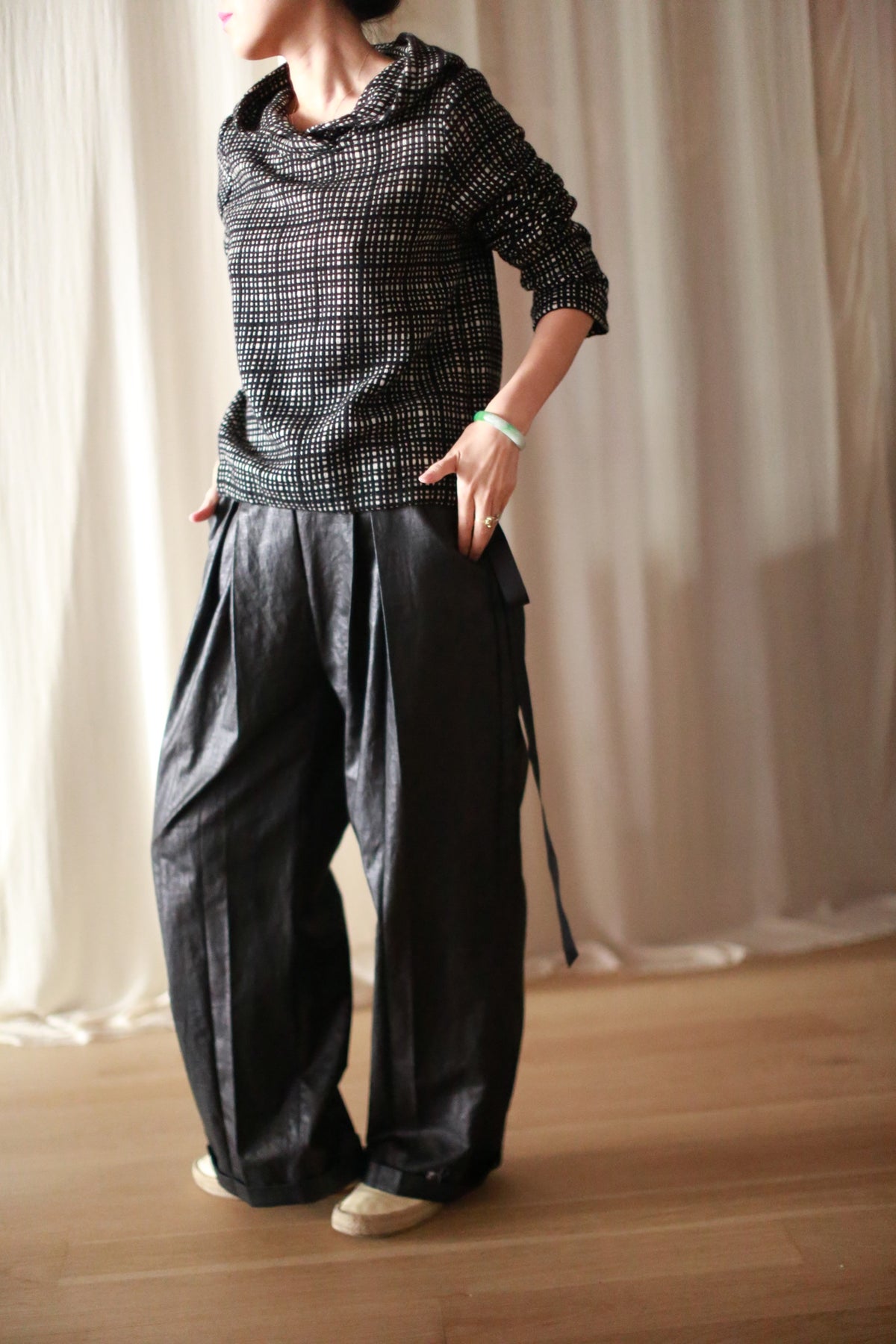 A person wears an Alex Hoodie in plaid, matched with loose, pleated dark trousers featuring a silk ribbon waist. They're indoors on a wooden floor with their hands in their pockets against a white curtain backdrop. Their face is not visible.