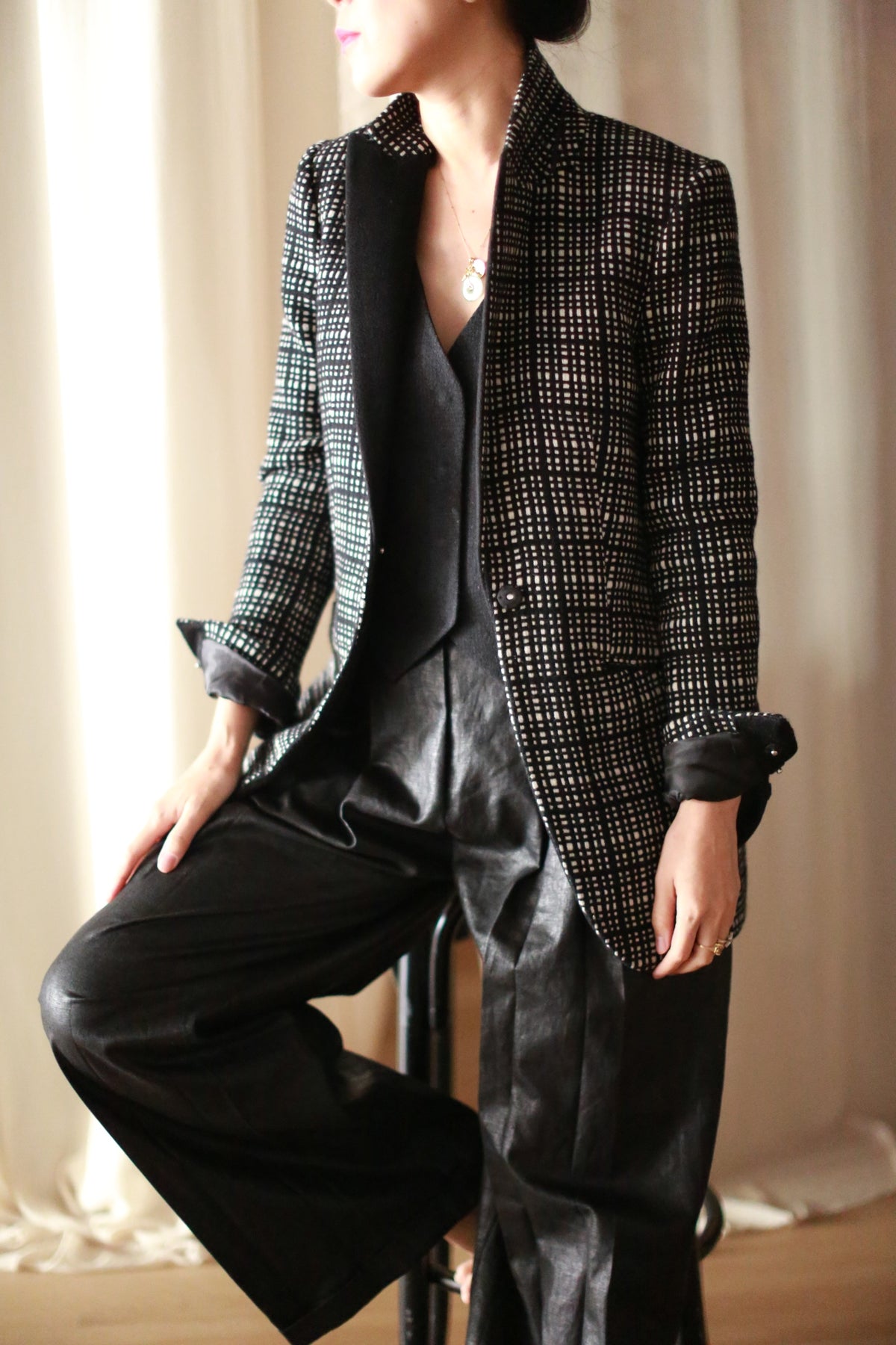 A person sits on a stool wearing the Sloane | Plaid tuxedo jacket over a black top and wide-leg black pants, set against light-colored curtains. The person's face is turned away and not fully visible.