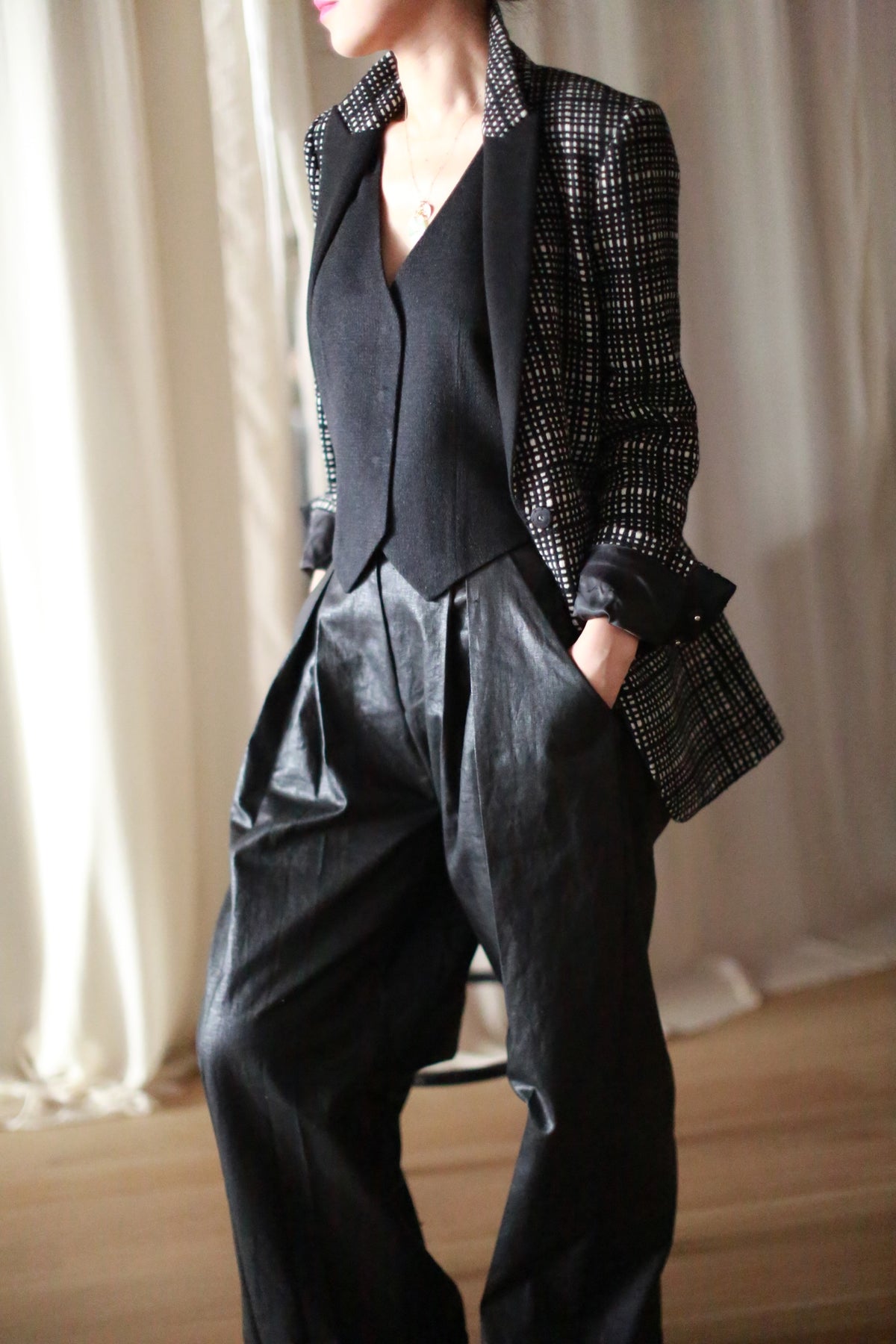 A person wears the Sloane | Plaid black cashmere blazer, a black vest, and wide-leg black trousers, standing with hands in pockets against a softly lit neutral background. The person's face is partially out of frame.