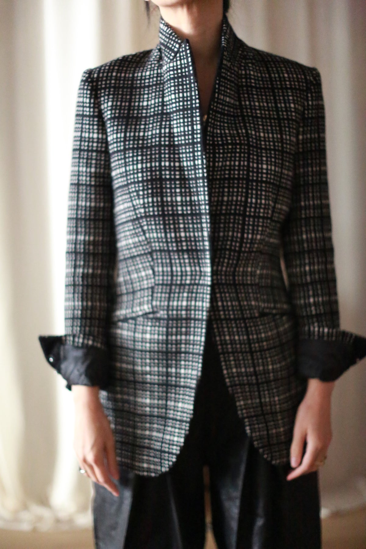 A person wears the Sloane | Plaid black and white cashmere blazer with rolled-up sleeves and dark pants, standing indoors against a blurred, light background. Their face is partially out of frame.
