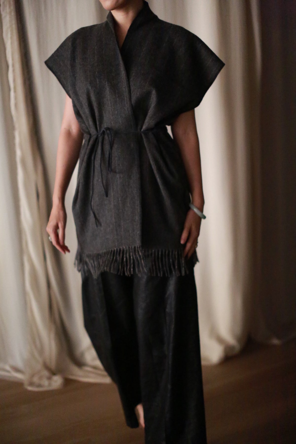 A person in a dark, loose outfit with wide sleeves, a waist tie, and fringe at the hem stands indoors by draped curtains, styled with the Men's Scarf Vest | Charcoal.