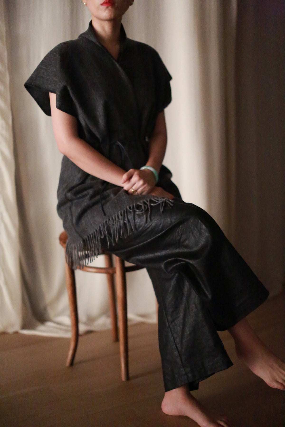 A person with short hair sits barefoot on a wooden chair, wearing the Men's Scarf Vest in Charcoal. Their loose pants add comfort, hands folded on their lap, and a light curtain forms the background. Their face is partially visible.